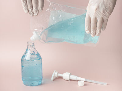 Hands With Gloves Refill Blue Alcohol Gel Into Plastic Bottle. Pink Pastel Background.