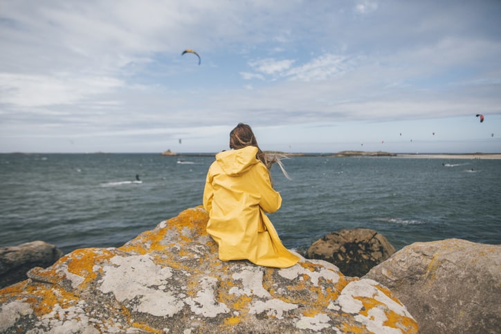 Junge Frau mit Regenjacke aus Kunststoff sitz an Küste - Klimaschutz