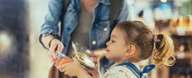 Nahaufnahme eines kleinen Mädchens, das mit einem Rucksack im Einkaufswagen sitzt. Sie hat Haare zu einem Pferdeschwanz gebunden. Sie hält Plastikbehälter mit Lebensmitteln aus einem Kühlschrank, den ihre nicht wiederzuerkennende Mutter gepflückt hat.