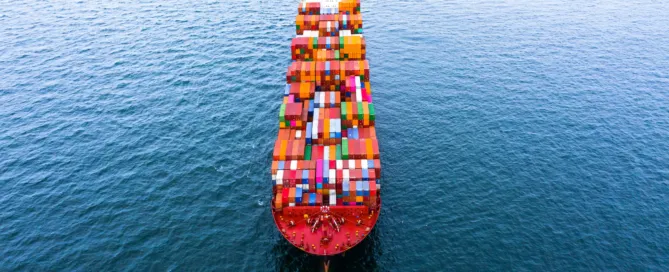 Luftaufnahme eines großen Containerschiffs, das mit bunten Frachtcontainern beladen über dunkelblaues Meer fährt.