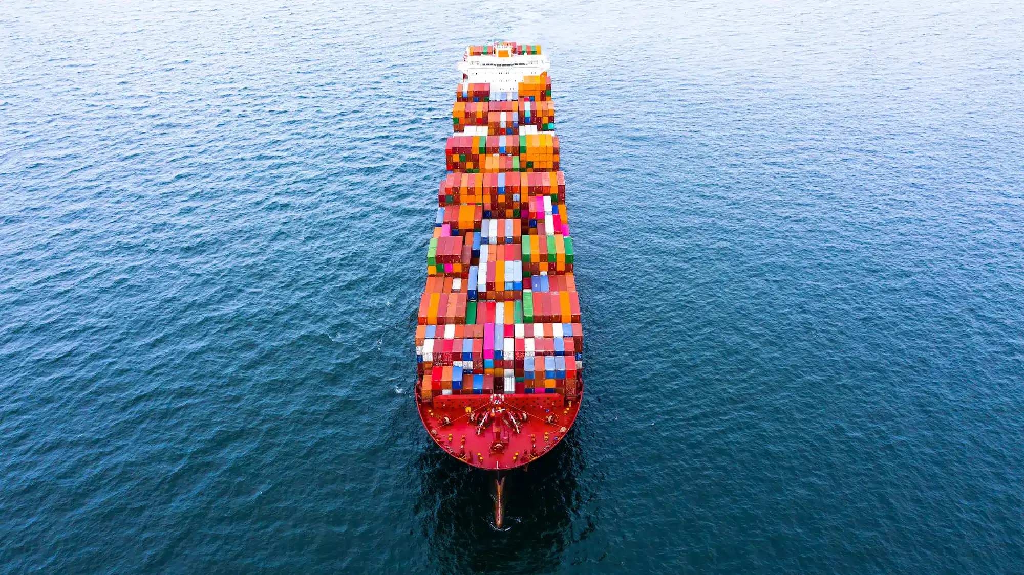 Containerschiff auf offenem Meer Luftaufnahme eines großen Containerschiffs, das mit bunten Frachtcontainern beladen über dunkelblaues Meer fährt.